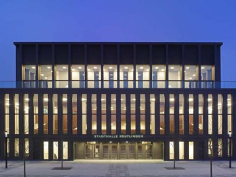 Ein modernes, rechteckiges Gebäude mit großen vertikalen Fenstern wird in der Abenddämmerung von innen beleuchtet. Auf dem Eingang steht "Stadthalle Reutlingen".