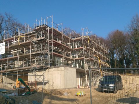 Ein mehrstöckiges, im Bau befindliches Gebäude mit Gerüsten auf allen Seiten, Baumaterialien auf dem Boden und einem schwarzen Lieferwagen, der in der Nähe parkt. Bäume und blauer Himmel im Hintergrund.