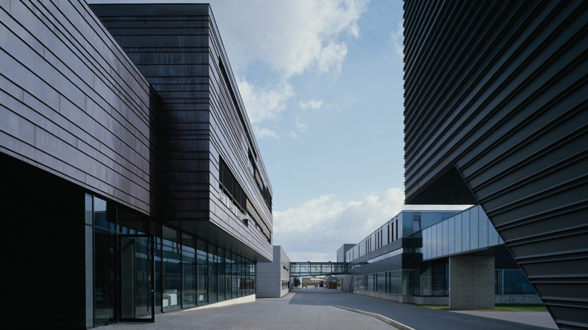 Moderne Bürogebäude mit dunklen Metallfassaden und Glasfenstern säumen einen gepflasterten Fußgängerweg unter einem teilweise bewölkten Himmel.