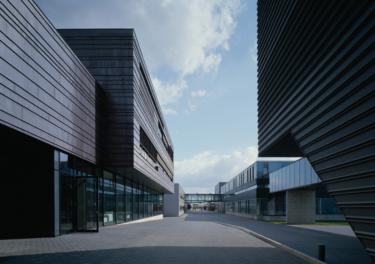 Moderne Bürogebäude mit dunklen Metallfassaden und Glasfenstern säumen einen gepflasterten Fußgängerweg unter einem teilweise bewölkten Himmel.