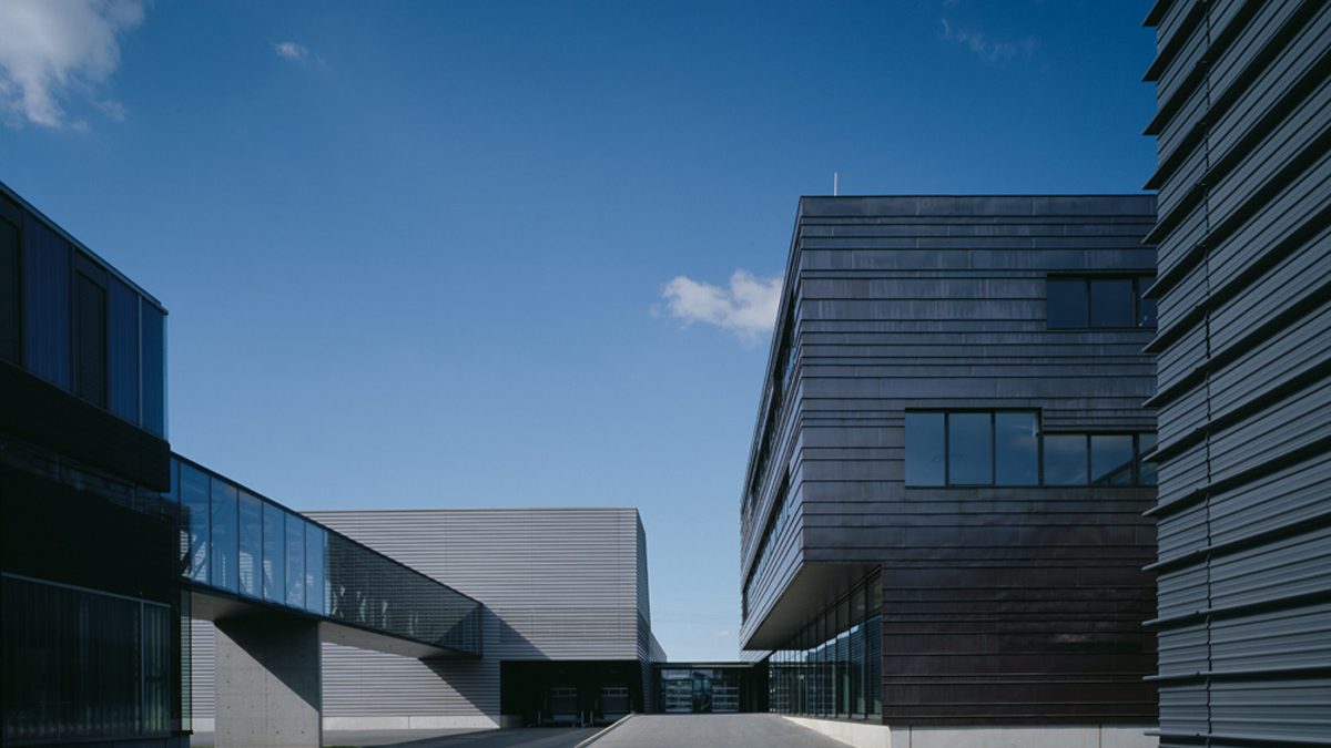 Moderne Industriegebäude mit Metallfassaden und großen Fenstern unter strahlend blauem Himmel, verbunden durch einen gläsernen Gang, mit einem leeren gepflasterten Hof im Vordergrund.