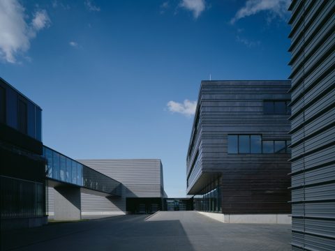 Moderne Industriegebäude mit Metallfassaden und großen Fenstern unter strahlend blauem Himmel, verbunden durch einen gläsernen Gang, mit einem leeren gepflasterten Hof im Vordergrund.