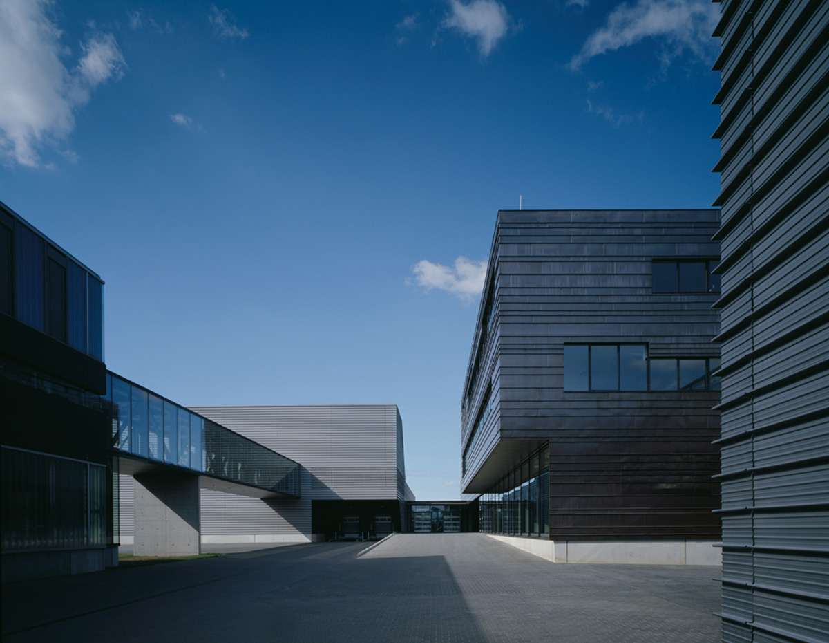 Moderne Industriegebäude mit Metallfassaden und großen Fenstern unter strahlend blauem Himmel, verbunden durch einen gläsernen Gang, mit einem leeren gepflasterten Hof im Vordergrund.