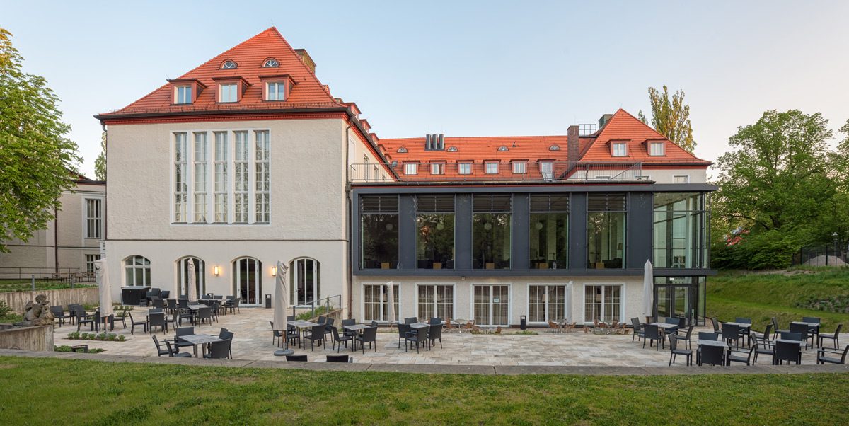 Dreistöckiges Gebäude mit roten Ziegeldächern, großen Fenstern und einem Glasanbau; Außenterrasse mit schwarzen Tischen und Stühlen auf Steinpflaster.