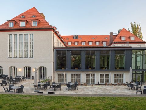 Dreistöckiges Gebäude mit roten Ziegeldächern, großen Fenstern und einem Glasanbau; Außenterrasse mit schwarzen Tischen und Stühlen auf Steinpflaster.