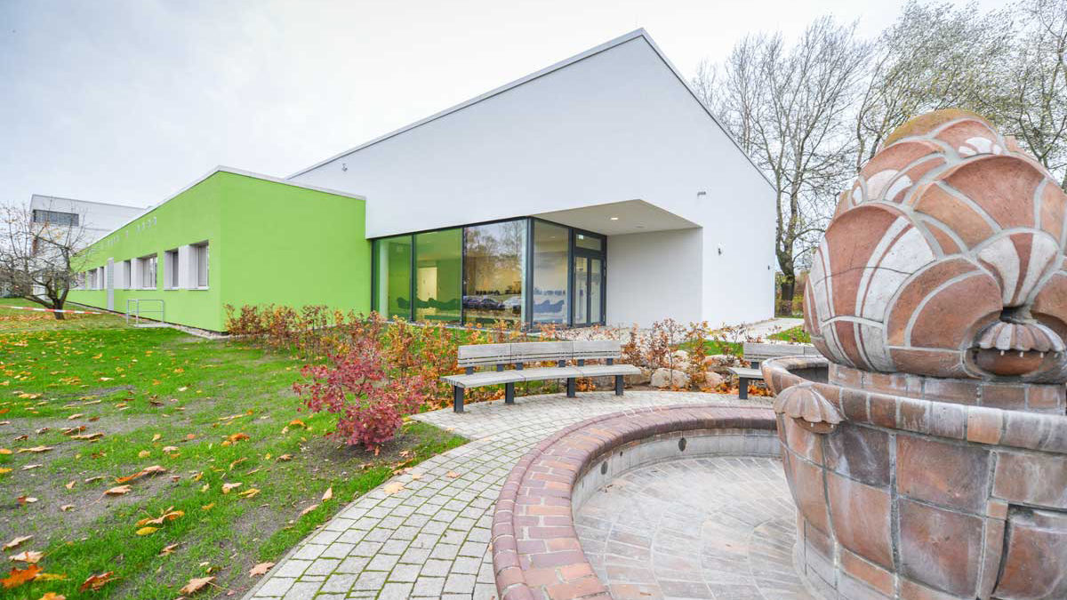 Modernes Gebäude mit weißem und grünem Äußeren, großen Fenstern, umgeben von Herbstlaub, mit einem Steinbrunnen und Bänken im Vordergrund.