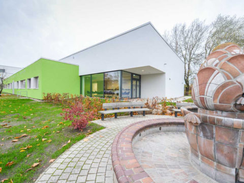 Modernes Gebäude mit weißem und grünem Äußeren, großen Fenstern, umgeben von Herbstlaub, mit einem Steinbrunnen und Bänken im Vordergrund.