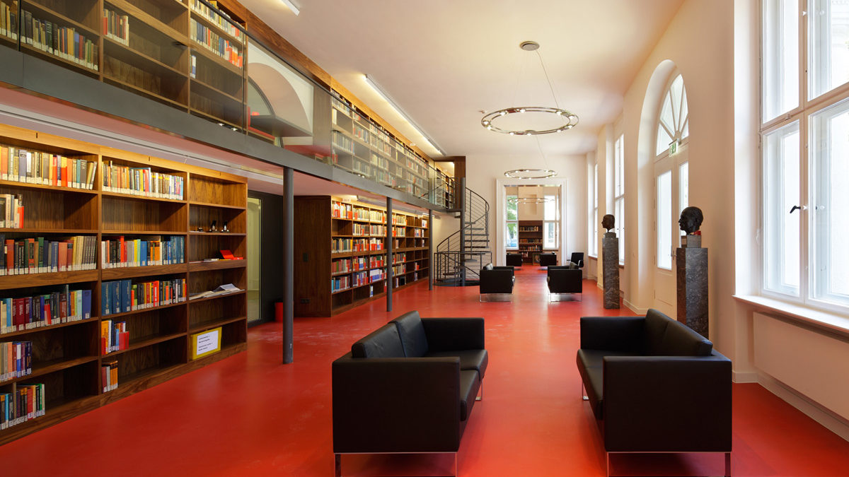 Geräumige Bibliothek mit rotem Boden, schwarzen Sofas, hohen Bücherregalen, großen Fenstern und einem Zwischengeschoss, das über eine Wendeltreppe erreichbar ist.
