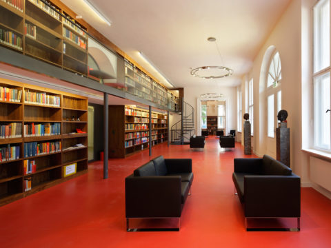 Geräumige Bibliothek mit rotem Boden, schwarzen Sofas, hohen Bücherregalen, großen Fenstern und einem Zwischengeschoss, das über eine Wendeltreppe erreichbar ist.