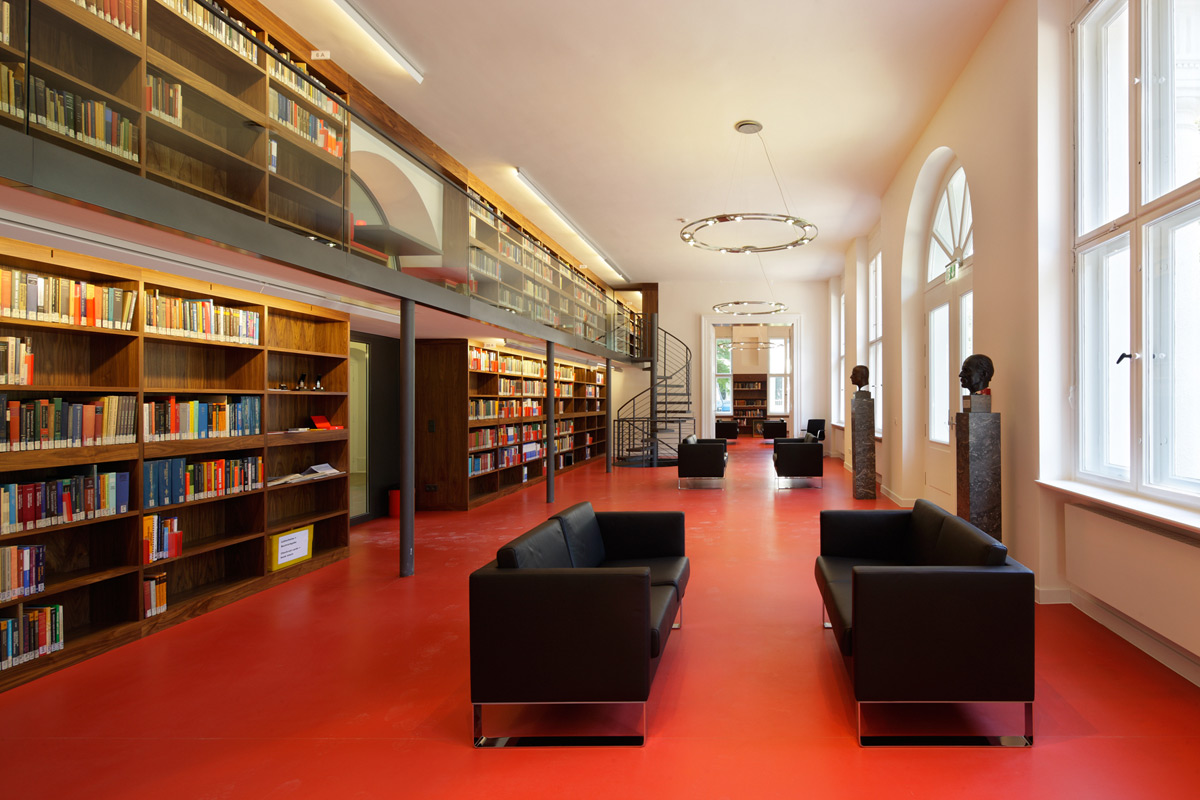 Geräumige Bibliothek mit rotem Boden, schwarzen Sofas, hohen Bücherregalen, großen Fenstern und einem Zwischengeschoss, das über eine Wendeltreppe erreichbar ist.