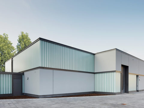 Modernes, minimalistisches Gebäude mit Milchglasscheiben und klaren geometrischen Linien, umgeben von Bäumen und einer gepflasterten Fläche, unter freiem Himmel.