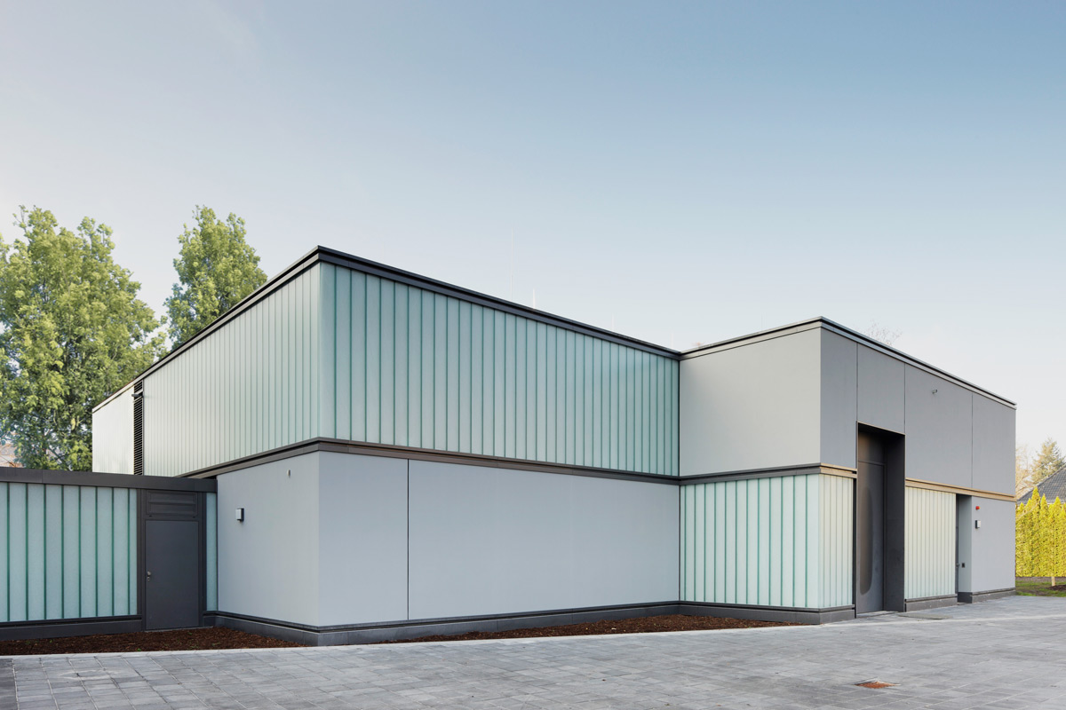 Modernes, minimalistisches Gebäude mit Milchglasscheiben und klaren geometrischen Linien, umgeben von Bäumen und einer gepflasterten Fläche, unter freiem Himmel.