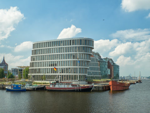 Ein modernes, geschwungenes Bürogebäude mit Glasfenstern steht am Wasser, vor dem mehrere Boote angedockt sind und über dem ein teilweise bewölkter Himmel hängt.