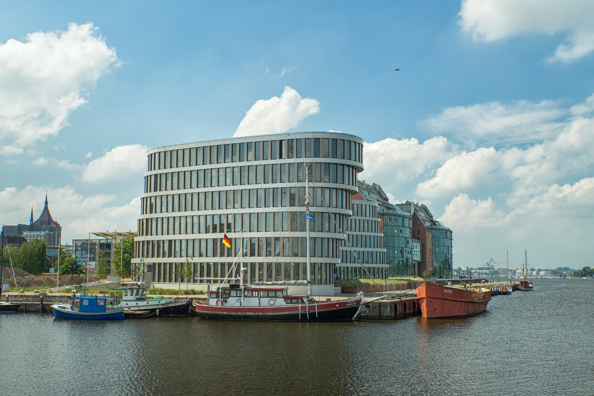 Ein modernes, geschwungenes Bürogebäude mit Glasfenstern steht am Wasser, vor dem mehrere Boote angedockt sind und über dem ein teilweise bewölkter Himmel hängt.