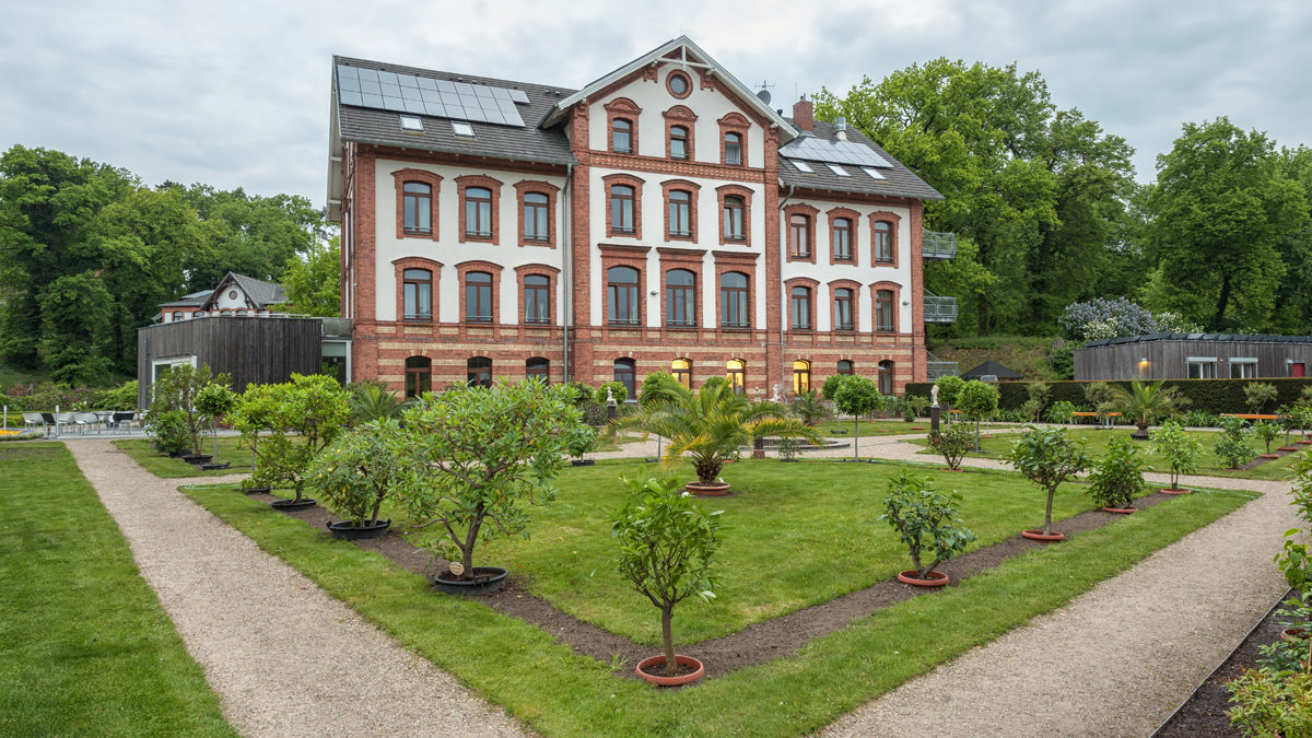 Ein dreistöckiges Backsteingebäude mit Sonnenkollektoren, umgeben von einem formalen Garten mit Kieswegen, gepflegtem Rasen und Topfpflanzen.