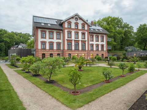 Ein dreistöckiges Backsteingebäude mit Sonnenkollektoren, umgeben von einem formalen Garten mit Kieswegen, gepflegtem Rasen und Topfpflanzen.