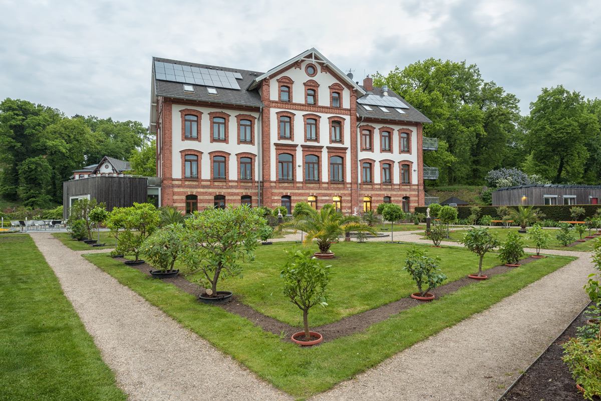 Ein dreistöckiges Backsteingebäude mit Sonnenkollektoren, umgeben von einem formalen Garten mit Kieswegen, gepflegtem Rasen und Topfpflanzen.