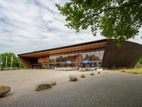 Ein modernes, geschwungenes Holzgebäude mit großen Glasfenstern beherbergt ein Café mit Sitzgelegenheiten im Freien; im Vordergrund befinden sich ein mit Steinen gepflasterter Platz und verstreute Felsen.