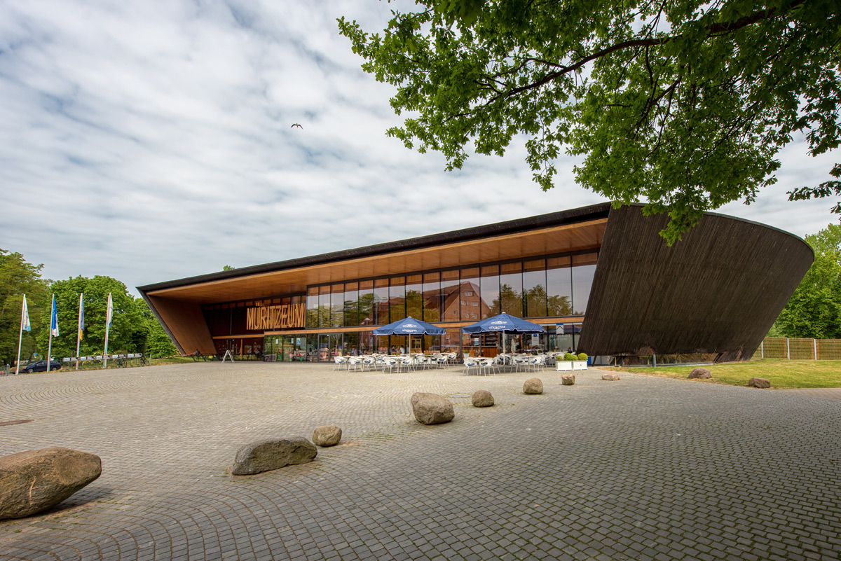 Ein modernes, geschwungenes Holzgebäude mit großen Glasfenstern beherbergt ein Café mit Sitzgelegenheiten im Freien; im Vordergrund befinden sich ein mit Steinen gepflasterter Platz und verstreute Felsen.