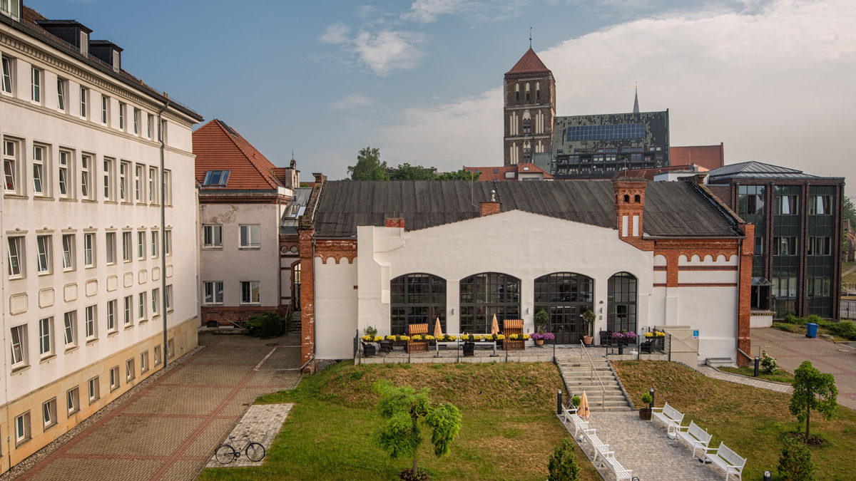 Ein weißes Gebäude mit großen Bogenfenstern und Sitzgelegenheiten im Freien befindet sich zwischen modernen und historischen Strukturen, mit Bänken und einer kleinen Rasenfläche davor. Im Hintergrund ist ein Kirchturm zu sehen.