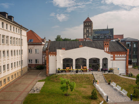 Ein weißes Gebäude mit großen Bogenfenstern und Sitzgelegenheiten im Freien befindet sich zwischen modernen und historischen Strukturen, mit Bänken und einer kleinen Rasenfläche davor. Im Hintergrund ist ein Kirchturm zu sehen.