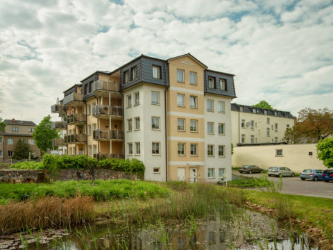 Ein vierstöckiges Mehrfamilienhaus mit Balkonen steht bei teilweise bewölktem Himmel in der Nähe eines kleinen Teichs mit Schilf. Im Hintergrund sind mehrere geparkte Autos und andere Gebäude zu sehen.
