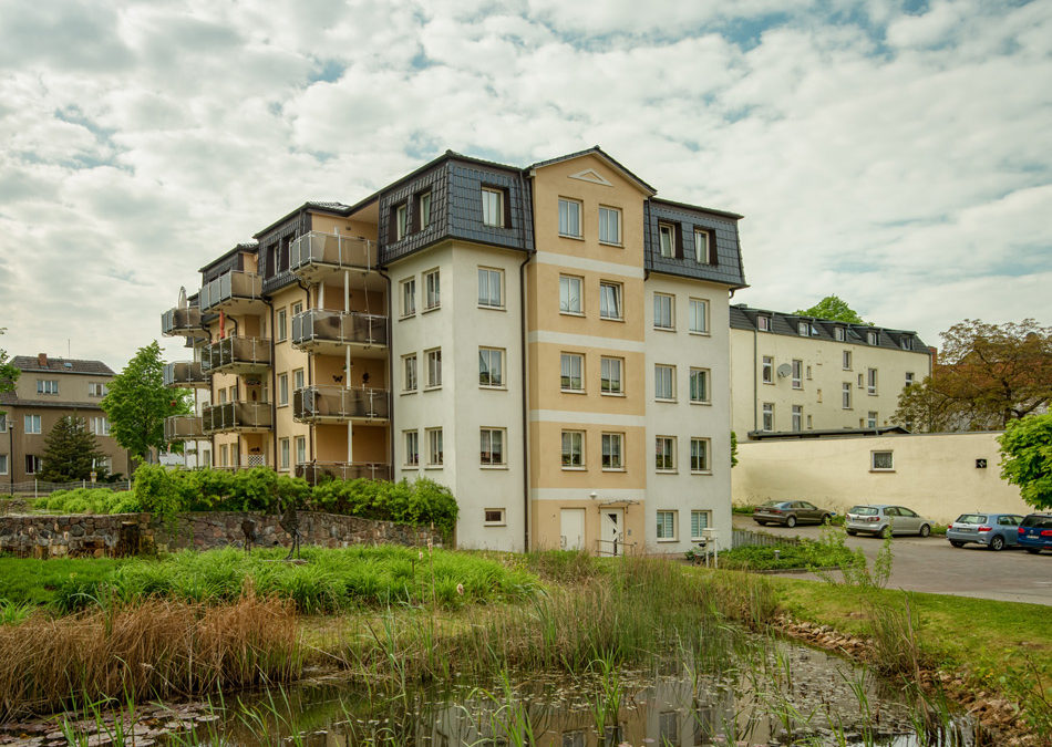 Ein vierstöckiges Mehrfamilienhaus mit Balkonen steht bei teilweise bewölktem Himmel in der Nähe eines kleinen Teichs mit Schilf. Im Hintergrund sind mehrere geparkte Autos und andere Gebäude zu sehen.