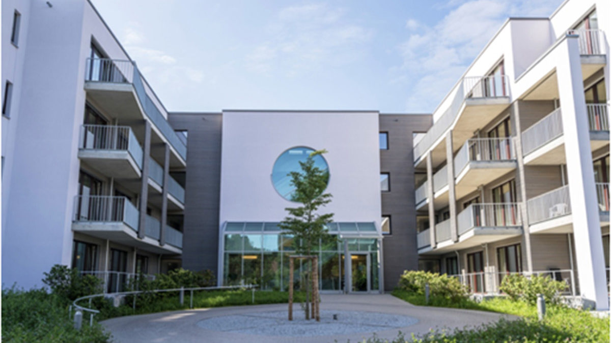 Modernes Mehrfamilienhaus mit Balkonen, einem zentralen Innenhof mit einem jungen Baum und einem gläsernen Eingang unter einem runden Fenster an einem sonnigen Tag.