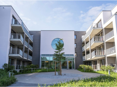 Modernes Mehrfamilienhaus mit Balkonen, einem zentralen Innenhof mit einem jungen Baum und einem gläsernen Eingang unter einem runden Fenster an einem sonnigen Tag.