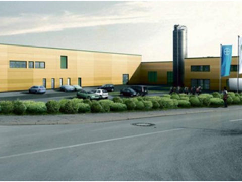 Ein modernes Industriegebäude mit gelben Wänden, geparkten Autos, grüner Bepflanzung und mehreren Fahnen am Eingang.