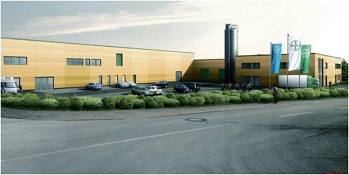 Ein modernes Industriegebäude mit gelben Wänden, geparkten Autos, grüner Bepflanzung und mehreren Fahnen am Eingang.