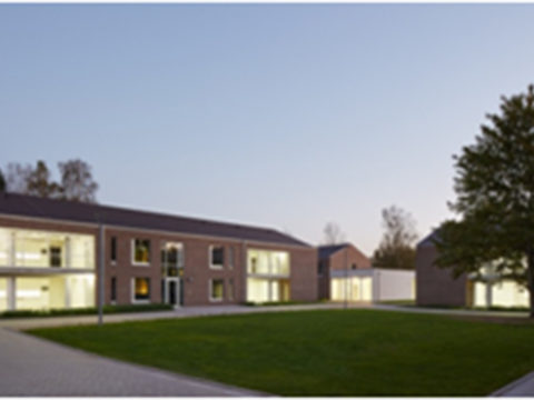 Ein modernes zweistöckiges Backsteingebäude mit großen, von innen beleuchteten Fenstern umgibt in der Abenddämmerung einen grünen Rasen.