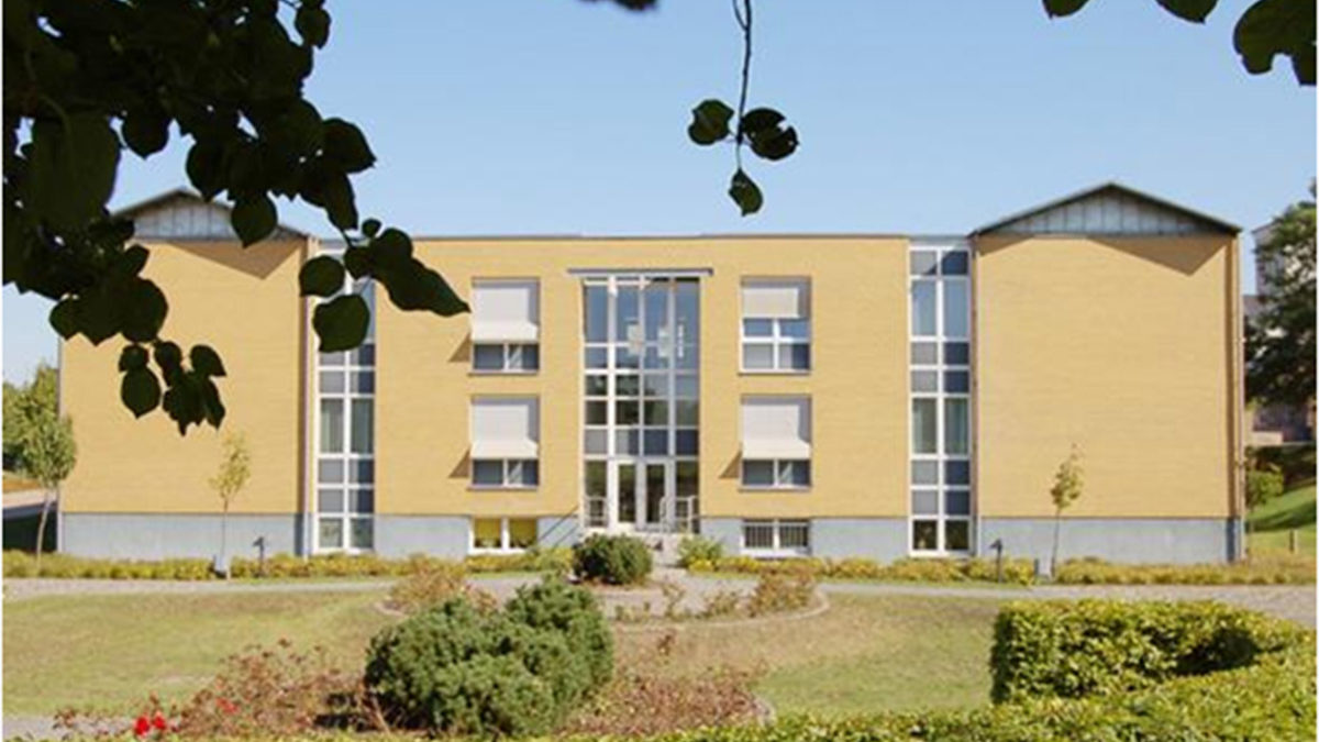 Ein modernes, rechteckiges, gelbes Backsteingebäude mit großen Glasfenstern, umgeben von gestutzten Hecken, Büschen und kleinen Bäumen unter einem klaren blauen Himmel.