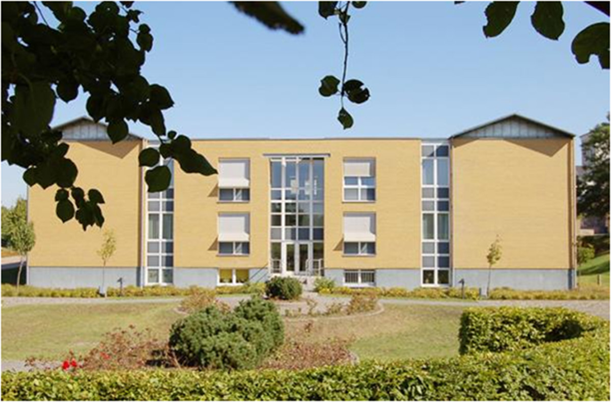 Ein modernes, rechteckiges, gelbes Backsteingebäude mit großen Glasfenstern, umgeben von gestutzten Hecken, Büschen und kleinen Bäumen unter einem klaren blauen Himmel.