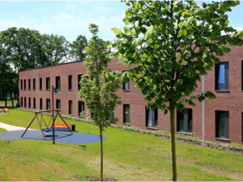 Ein modernes zweistöckiges Backsteingebäude mit großen Fenstern, umgeben von Gras, Bäumen und einem kleinen Spielplatz mit Schaukeln im Vordergrund.