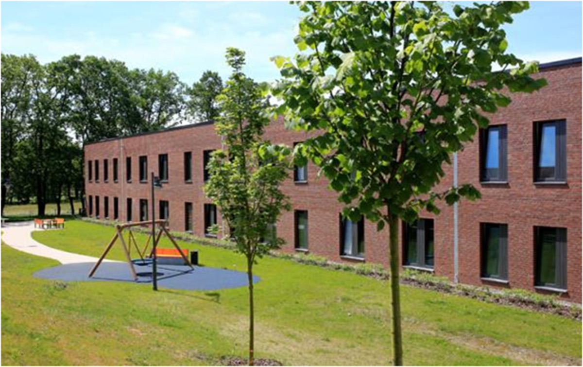 Ein modernes zweistöckiges Backsteingebäude mit großen Fenstern, umgeben von Gras, Bäumen und einem kleinen Spielplatz mit Schaukeln im Vordergrund.