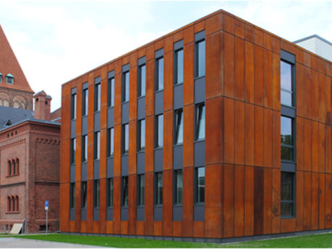 Ein modernes Gebäude mit einer rostfarbenen Fassade steht neben älteren Backsteinbauten mit Bogenfenstern und einem Turm.