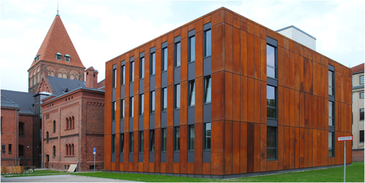 Ein modernes Gebäude mit einer rostfarbenen Fassade steht neben älteren Backsteinbauten mit Bogenfenstern und einem Turm.