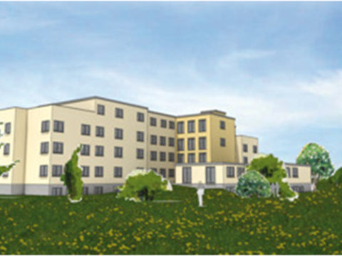 Ein vierstöckiges, beigefarbenes Gebäude mit vielen Fenstern steht in einem grasbewachsenen Feld mit Bäumen und blauem Himmel im Hintergrund.