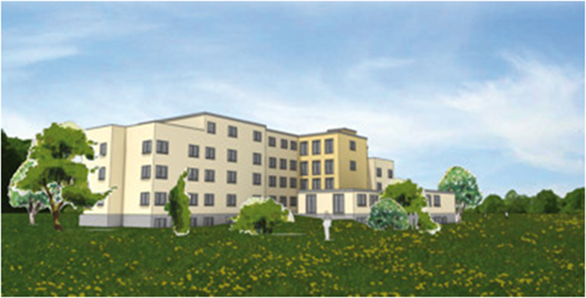 Ein vierstöckiges, beigefarbenes Gebäude mit vielen Fenstern steht in einem grasbewachsenen Feld mit Bäumen und blauem Himmel im Hintergrund.