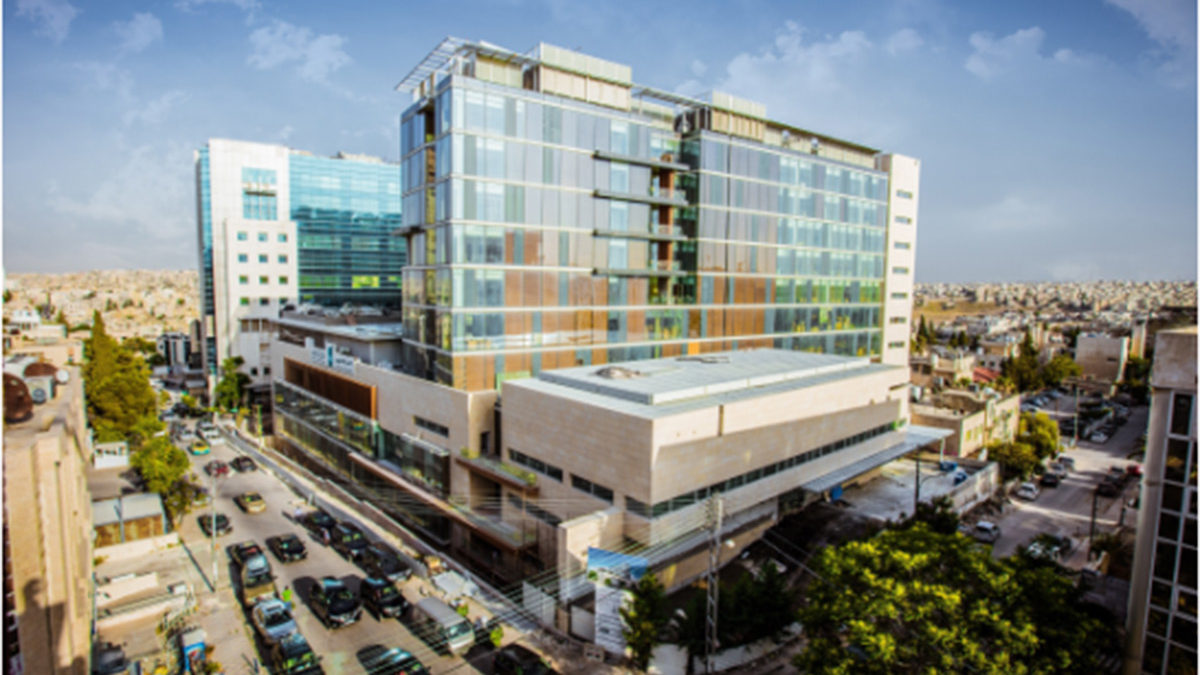 Ein modernes, mehrstöckiges Gebäude aus Glas und Stein steht an einer belebten Straßenkreuzung, umgeben von geparkten Autos und angrenzenden Gebäuden unter einem klaren Himmel.