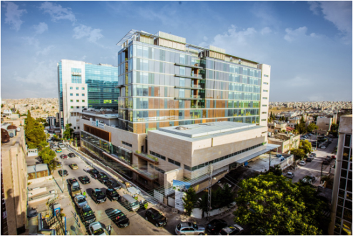 Ein modernes, mehrstöckiges Gebäude aus Glas und Stein steht an einer belebten Straßenkreuzung, umgeben von geparkten Autos und angrenzenden Gebäuden unter einem klaren Himmel.