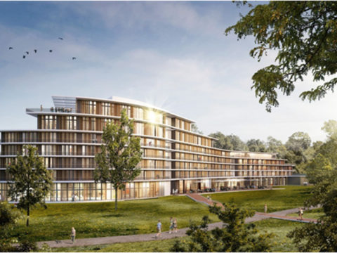 Ein modernes, mehrstöckiges Gebäude mit einer Fassade aus Glas und Holz steht in einem grasbewachsenen Park, umgeben von Bäumen und Menschen, die unter einem sonnigen Himmel auf den nahen Wegen spazieren gehen.