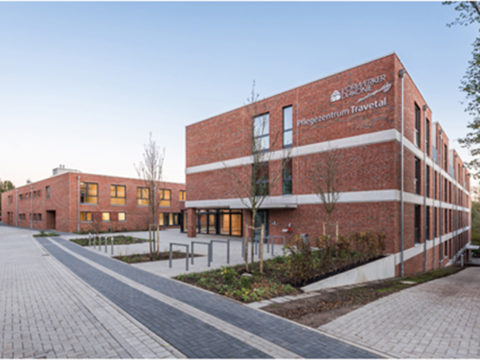 Ein modernes, dreistöckiges Backsteingebäude mit großen Fenstern und einem Schild mit der Aufschrift Pflegezentrum Travetal an der Fassade; ein weiteres ähnliches Gebäude befindet sich im Hintergrund.