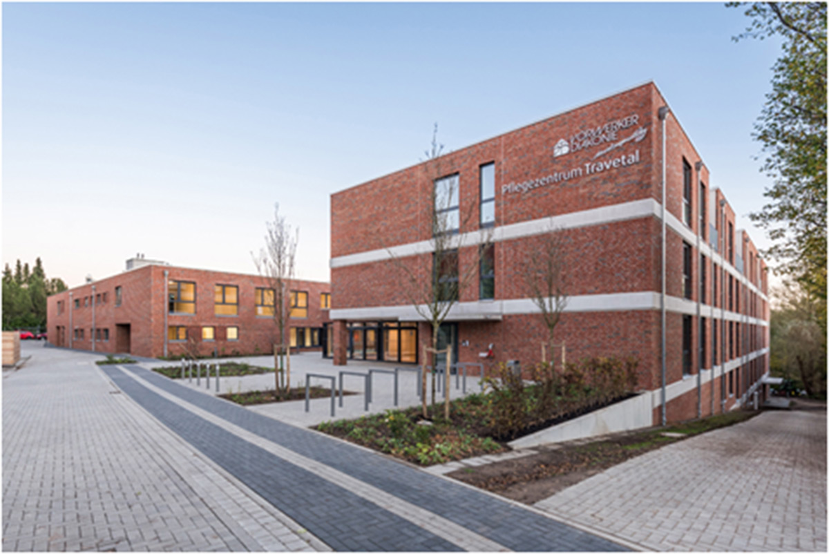 Ein modernes, dreistöckiges Backsteingebäude mit großen Fenstern und einem Schild mit der Aufschrift Pflegezentrum Travetal an der Fassade; ein weiteres ähnliches Gebäude befindet sich im Hintergrund.