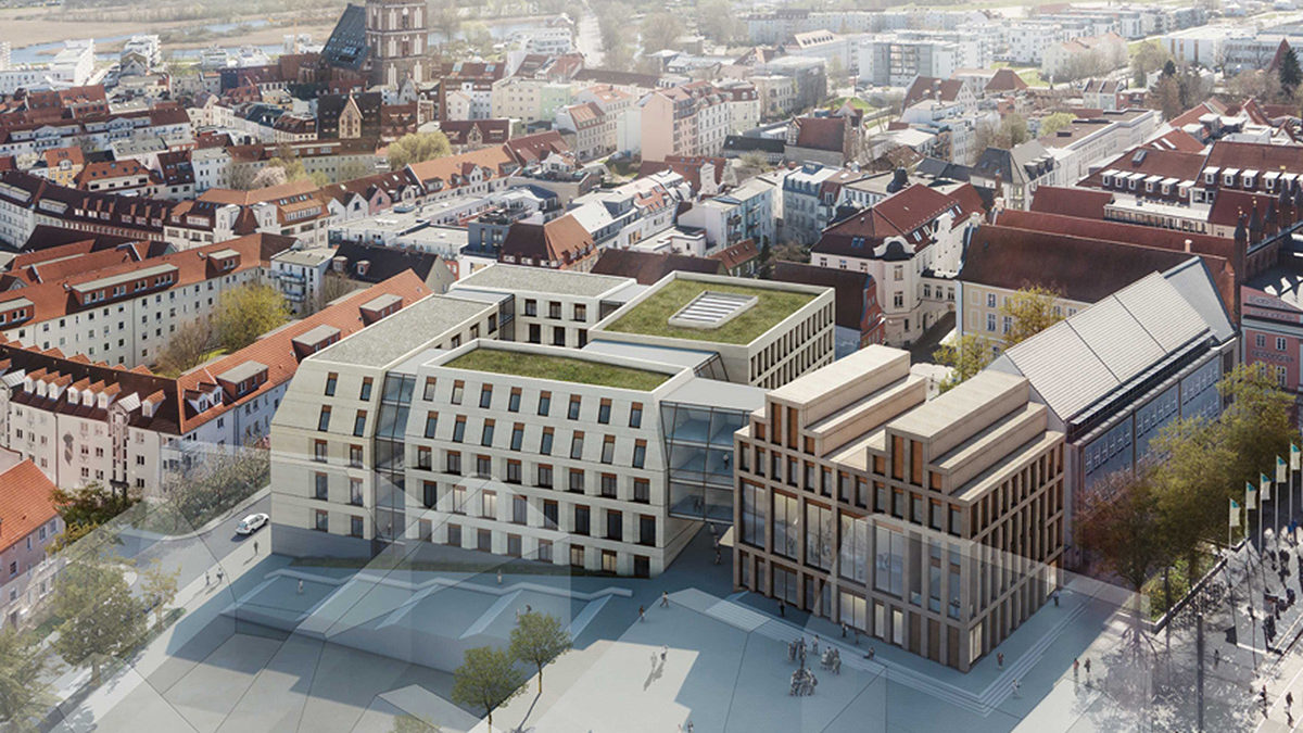 Luftaufnahme eines modernen, mehrstöckigen Gebäudekomplexes mit begrünten Dächern inmitten älterer, rot gedeckter Gebäude in einer europäischen Stadt.