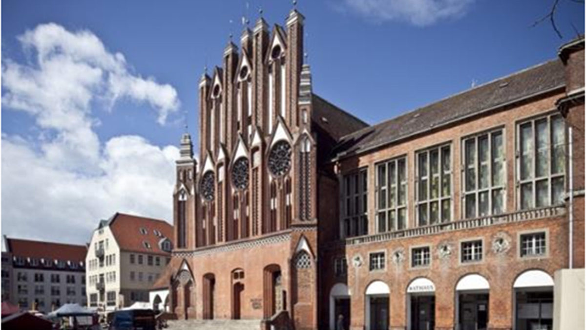 Ein historisches Gebäude der Backsteingotik mit verschnörkelten Bogenfenstern steht neben einem modernen Bau mit großen rechteckigen Fenstern auf einem europäischen Stadtplatz.