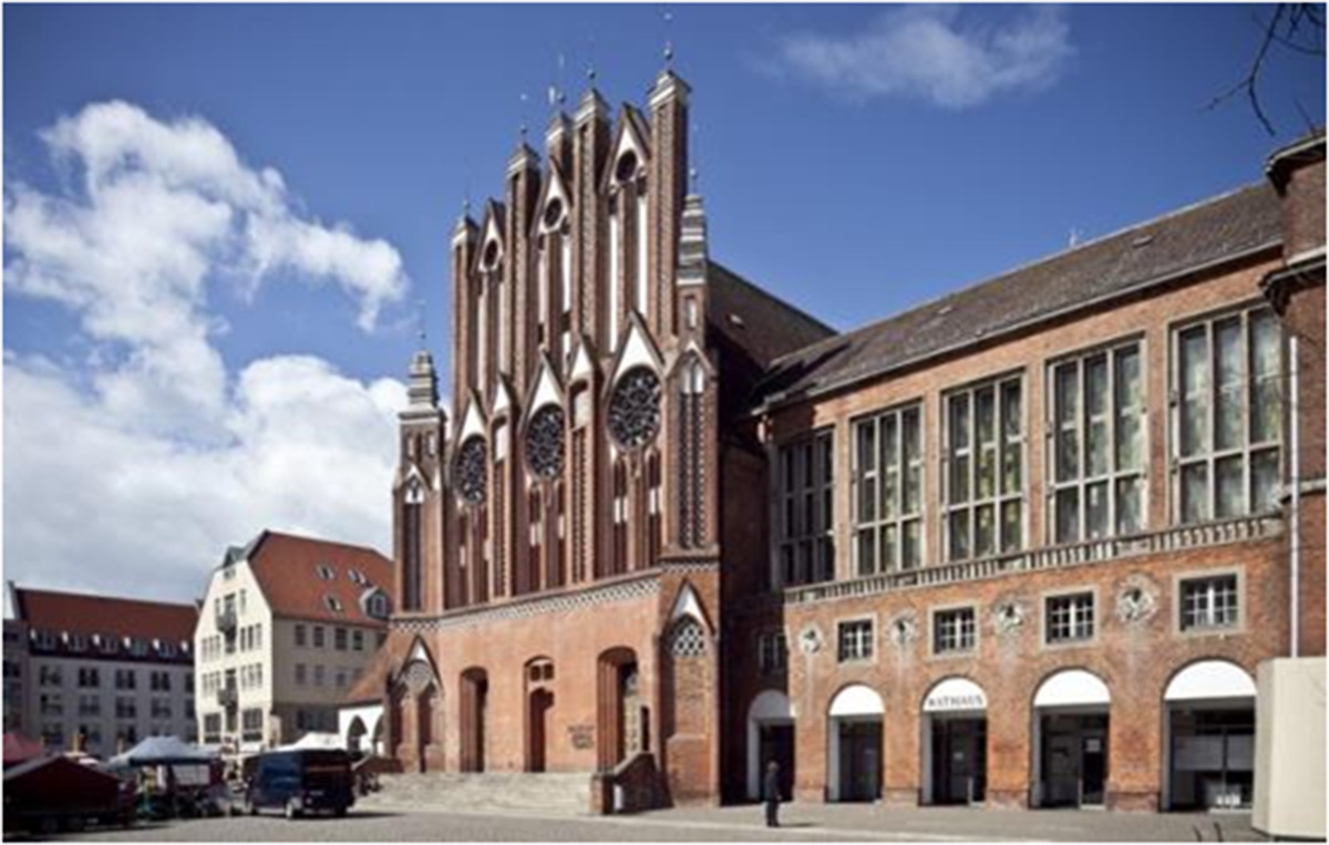 Ein historisches Gebäude der Backsteingotik mit verschnörkelten Bogenfenstern steht neben einem modernen Bau mit großen rechteckigen Fenstern auf einem europäischen Stadtplatz.