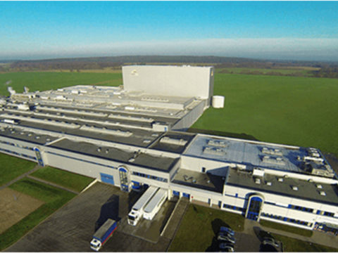 Luftaufnahme einer großen Industrieanlage mit mehreren Gebäuden, geparkten Fahrzeugen und angrenzenden grünen Feldern bei klarem Himmel.