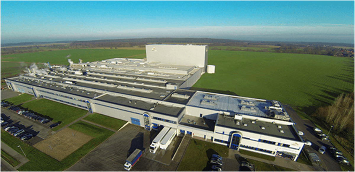 Luftaufnahme einer großen Industrieanlage mit mehreren Gebäuden, geparkten Fahrzeugen und angrenzenden grünen Feldern bei klarem Himmel.